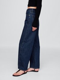 Weite, dunkelblaue Jeans mit hoher Taille, ausgestattet mit Fronttaschen und einer subtilen Waschung. Kombiniert mit schwarzen flachen Sandalen und einem taillierten schwarzen Oberteil.