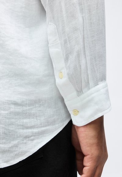 Κοντινό πλάνο ενός βραχίονα ατόμου που φοράει λευκό πουκάμισο από λινό με μακριά μανίκια, μανσέτα με κουμπιά και πλαϊνή σχισμή πάνω από μαύρο παντελόνι.