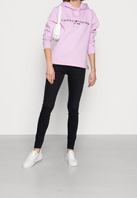 Rosa Kapuzenpullover mit Logo, schwarze enge Jeans und weiße Sneakers. Der Pullover hat eine Kordel und einen gerippten Saum. Einfaches, lässiges Design.