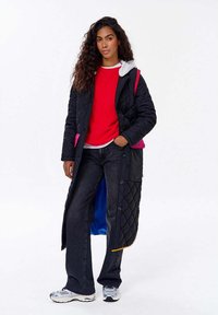 Žena s dlouhými kudrnatými vlasy, oblečená v dlouhém černém prošívaném kabátu, červeném svetru, �černých džínách a stříbrných teniskách, stojící s jednou rukou v kapse.