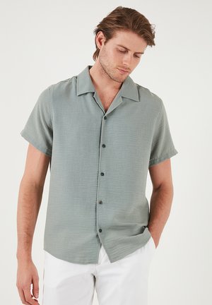 Camisa de manga corta verde claro con un ajuste relajado, que presenta un cuello con solapa y cinco botones oscuros en el frente. Textura suave.