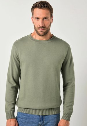 Man met kort bruin haar en baard, gekleed in een olijfgroene trui en blauwe spijkerbroek, staand tegen een effen lichte achtergrond.