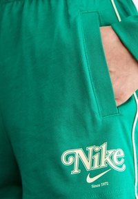 Zelené bavlněné šortky s boční kapsou, bílým lemováním a logem Nike světle krémové barvy s nápisem "Since 1972" pod ním.
