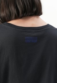 Close-up of a person’s back with short dark hair wearing a black shirt labeled "AMERICAN VINTAGE" near the neckline.