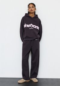 Tânără stă sprijinită de perete, purtând un hanorac negru supradimensionat cu textul "ninetyone" și pantaloni largi asortați, adidași bej pe un podea întunecată.