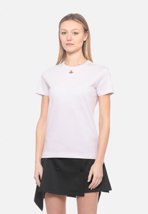 Donna con capelli biondi lisci che indossa una maglietta rosa chiaro tinta unita con un piccolo logo e una gonna nera asimmetrica, in piedi contro uno sfondo bianco.