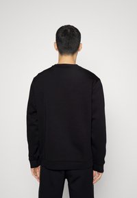 Man som står med ryggen mot kameran, iklädd en enkel svart långärmad sweatshirt och svarta byxor, mot en neutral ljus bakgrund.