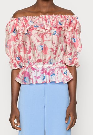 Persona que lleva una blusa rosa floral de hombros descubiertos con volantes y mangas abullonadas combinada con pantalones azul claro sobre un fondo liso.