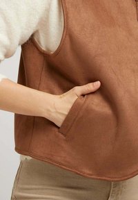 Main dans la poche d'un gilet sans manches en suède brun, porté par-dessus un pull blanc en peluche et un pantalon beige.