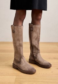 Bottines en daim marron jusqu'au genou avec une semelle plate, dotées de fermetures éclair sur les côtés et de détails de couture subtils, sur un fond de sol en bois.