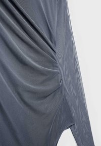 Tmavošedá priesvitná sieťovina zavesená a zhromaždená, aby vytvorila záhyby a textúru proti jednoduchému svetlému pozadiu.