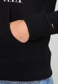 Svart sweatshirt med ribbad mudd, som har en framficka och en liten trefärgad logotyp. Texten på framsidan säger "D.E.N.I.M."