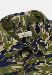 Chemise à boutons camouflage présentant une combinaison de motifs verts, bleus marine et beiges. Comprend un col et une étiquette avec le nom de la marque visible.