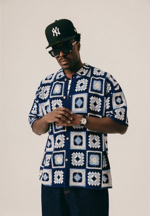 Mężczyzna w niebiesko-białej koszuli zrobionej na szydełku w kwadratowy wzór, czapce New York Yankees w kolorze czarnym, ciemnych okularach przeciwsłonecznych i zegarku na nadgarstku.