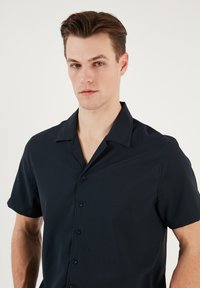Rövid ujjú, galléros ing sötét tengerészkék anyagból, sima felülettel és hat elülső gombbal. Egyszerű, mintázat nélküli dizájn jellemzi.