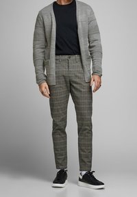 Cardigan grigio a trama sopra una maglietta nera, abbinato a pantaloni slim-fit a quadri in tonalità di marrone e verde, e sneakers nere con suole bianche.