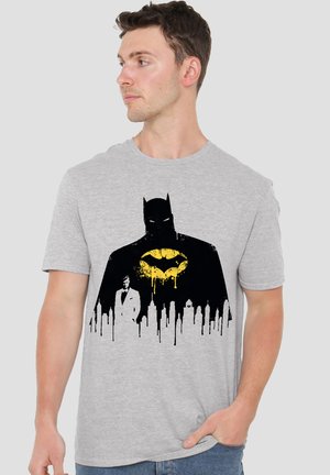 Mann trägt graues T-Shirt mit schwarzer Batman-Silhouette, gelbem Fledermaussymbol, Stadtsilhouette und Grafik eines Mannes im weißen Anzug, schaut nach links.