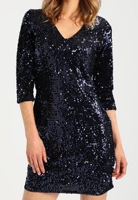 Abito blu navy con paillettes, scollo a V, maniche a tre quarti e silhouette aderente, caratterizzato da una superficie scintillante e strutturata.