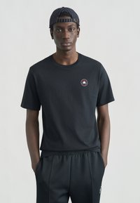 Μαύρο βαμβακερό T-shirt με στρογγυλή λαιμόκοψη, κοντά μανίκια και μικρό κυκλικό λογότυπο με αστέρι στην αριστερή πλευρά. Το μοντέλο φοράει παντελόνια που ταιριάζουν.