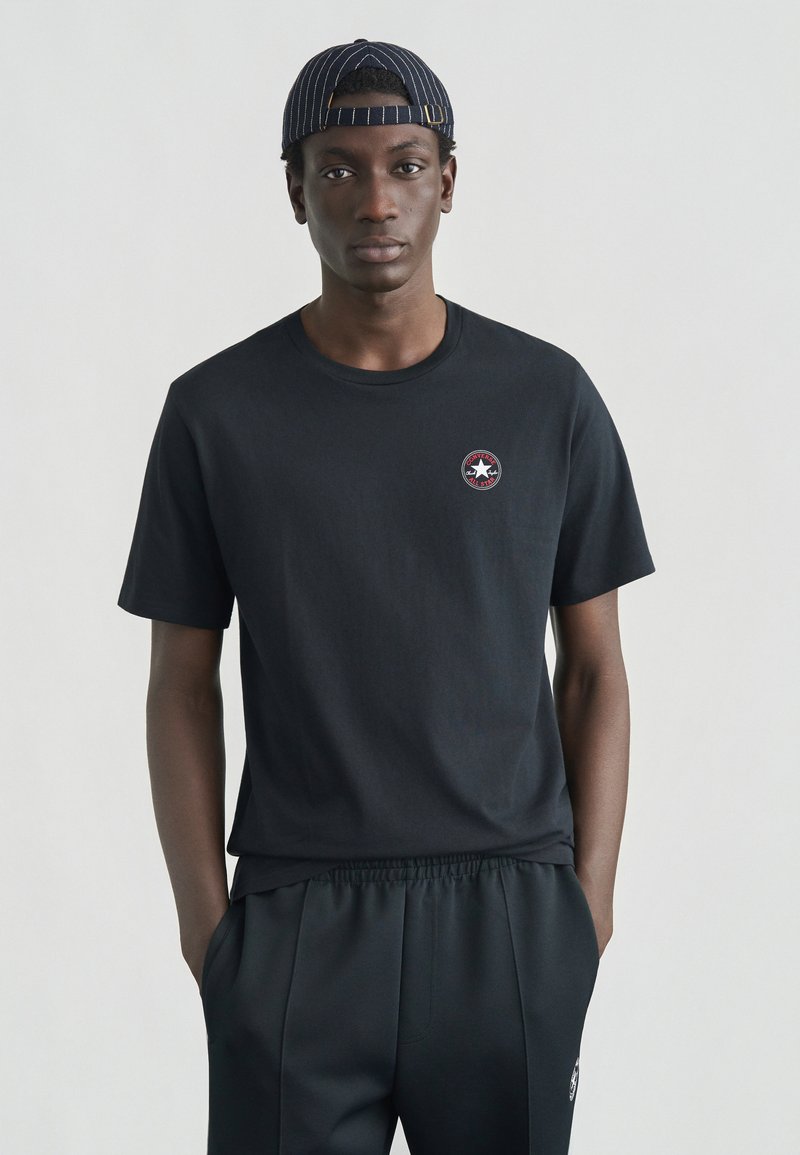 Μαύρο βαμβακερό T-shirt με στρογγυλή λαιμόκοψη, κοντά μανίκια και μικρό κυκλικό λογότυπο με αστέρι στην αριστερή πλευρά. Το μοντέλο φοράει παντελόνια που ταιριάζουν.