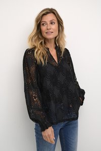 Črna čipkasta bluzka s cvetličnimi vzorci, V izrezom in dolgimi rokavi, ki imajo naborke na manšetah; kombinirana z modrimi jeans hlačami.