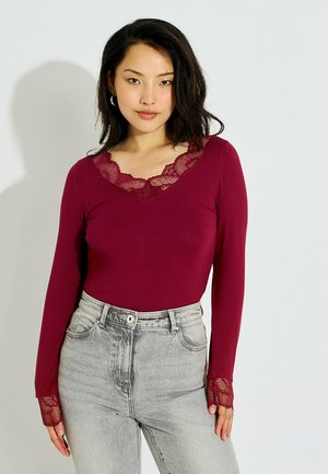 Mujer con una camiseta ajustada de manga larga burdeos con encaje en el escote y los puños, combinada con vaqueros de tiro alto en gris claro.