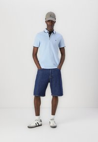 Lysblå polo-shirt med navy krave, denim shorts, hvide og sorte sneakers, og en grå kasket. Model står med hænderne i lommerne.