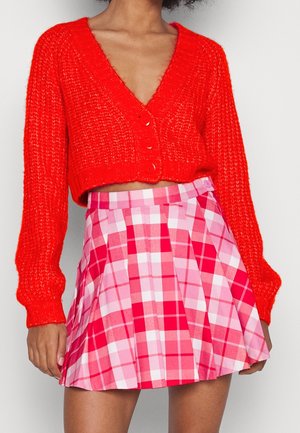 Femme portant un cardigan tricoté court rouge vif avec des boutons et une jupe plissée à carreaux roses et rouges taille haute.