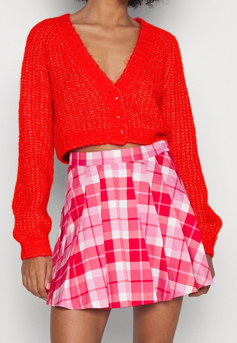 Femme portant un cardigan tricoté court rouge vif avec des boutons et une jupe plissée à carreaux roses et rouges taille haute.