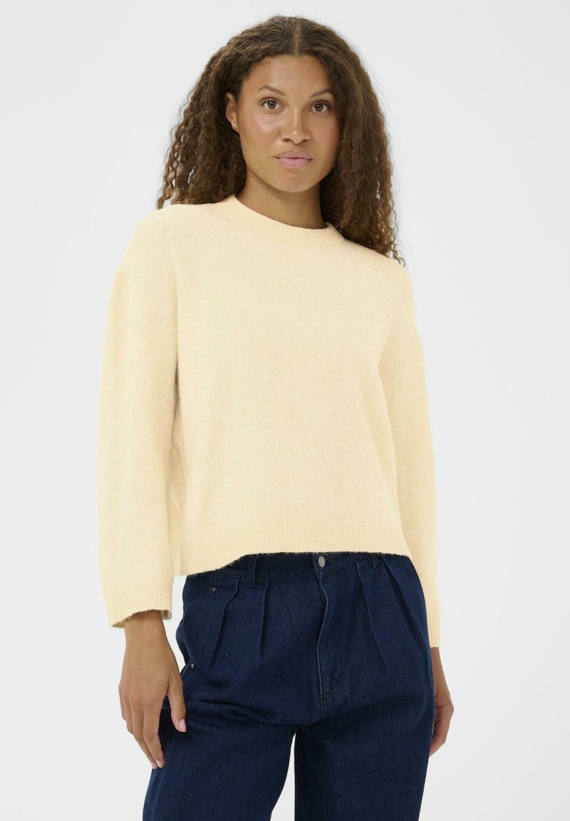 Pull cropped jaune claire avec un décolleté rond et une coupe ample. Tissu texturé, porté sur un pantalon en denim plissé bleu taille haute.
