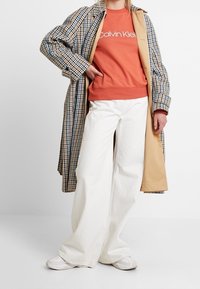 Lungo cappotto a quadri in blu e beige sopra una felpa arancione con scritte bianche, abbinato a pantaloni larghi bianchi e sneakers bianche.