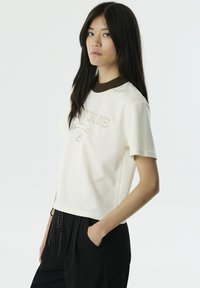 Lichtgekleurde T-shirt met een bruine ribbelkraag, voorzien van geborduurde tekst "UNVERSE" en decoratieve accenten. Korte mouwen en een relaxte pasvorm.