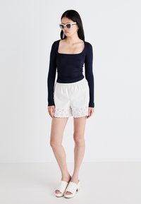 Top bleu marine à manches longues, short blanc avec des détails en œillets et mules ouvertes beiges. Tissu lisse avec une silhouette ajustée et une taille élastique.