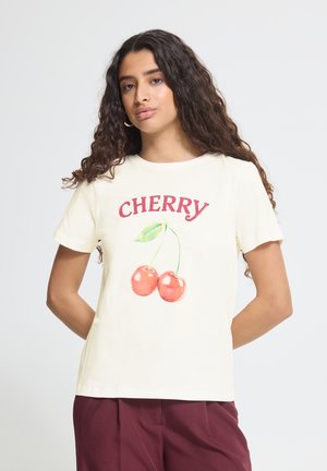 Jeune femme aux longs cheveux bouclés porte un T-shirt crème avec « CHERRY » et deux cerises imprimées sur le devant, assorti d’un pantalon bordeaux.