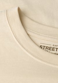 Bežové tričko s kulatým výstřihem, viditelně prošité na límci, s částečně potisknutou etiketou s nápisem "STREET."