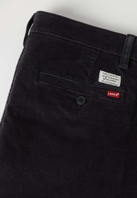 Pantalon en velours côtelé foncé avec une poche passepoilée arrière boutonnée et des étiquettes de la marque Levi's au-dessus de la poche.