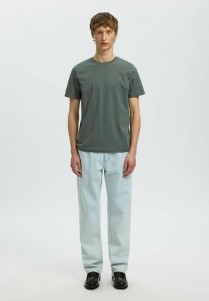 Ung mand stående, iført en mørkegrøn T-shirt, lyseblå jeans og sorte sko, mod en ensfarvet lys baggrund.