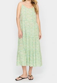Kobieta w sukience bez rękawów, sięgającej do kostek, w jasnym zielonym kolorze z kwiatowym wzorem, z cienkimi ramiączkami i czarnymi klapkami, stojąca na jednolitym tle.
