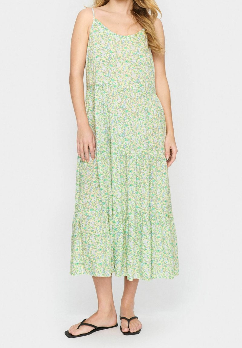 Kobieta w sukience bez rękawów, sięgającej do kostek, w jasnym zielonym kolorze z kwiatowym wzorem, z cienkimi ramiączkami i czarnymi klapkami, stojąca na jednolitym tle.