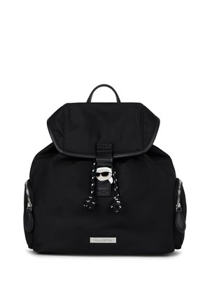 Musta reppu, jossa on läppä, etupuolen nyöri, sivutaskut vetoketjulla ja hopeinen logo-levy, jossa on klipsi, jossa on tyylitelty kasvokuva aurinkolaseja käyttäen.