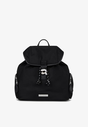Czarna plecak z klapą, przednim sznurkiem, bocznymi kieszeniami na zamek i srebrną tabliczką z logo z zapięciem przedstawiającą stylizowaną twarz w okularach przeciwsłonecznych.