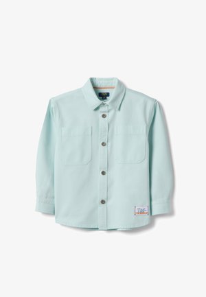 Camisa de manga larga azul claro con botones y dos bolsillos en el pecho, con un pequeño parche que dice "TAG THE WORLD" cerca del dobladillo.