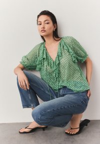 Green, sheer blouse with polka dots and ruffled neckline, paired with blue denim jeans and black flip-flops. Casual outfit.