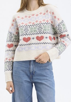 Kobieta w kremowym swetrze z czerwonymi sercami, zielonymi śnieżynkami i geometrycznymi wzorami, zestawionym z jasnoniebieskimi dżinsami.
