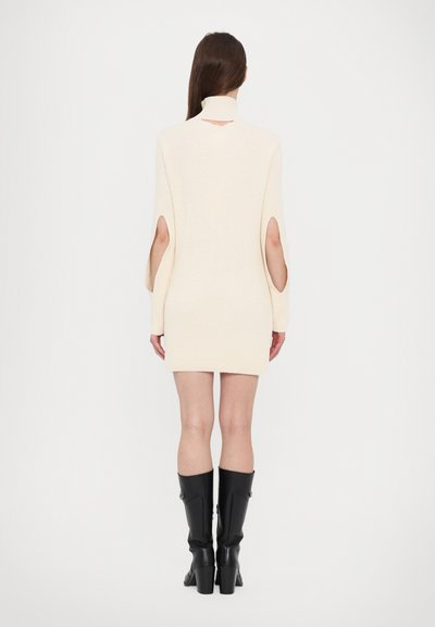 Rochie mini tricotată crem cu guler înalt, având decupaje la mâneci. Purtată cu cizme negre cu toc înalt. Textură netedă.