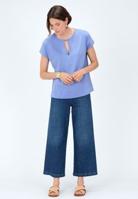 Lila kortef mouwen top met een keyhole halslijn, gecombineerd met wijde donkere spijkerbroek. Het model draagt geweven tan sandaaltjes en een gouden armband.