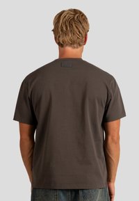 Camiseta de algodón marrón con cuello redondo, mangas cortas y una etiqueta rectangular en la parte posterior. Textura suave, corte relajado.