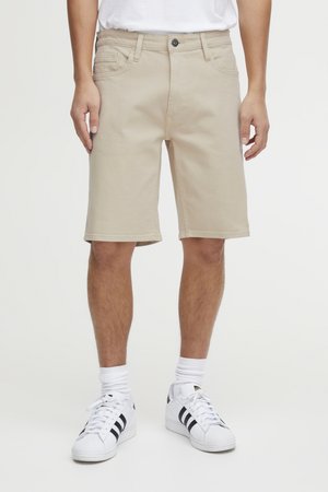 Modèle portant un short beige jusqu’aux genoux, une chemise blanche, des chaussettes blanches et des baskets blanches à rayures noires.