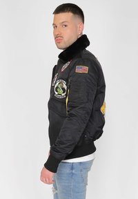 Svart bomberjacka med fuskpälskrage, prydd med flera märken, dragkedjor och ribbade manschetter. Bärs över en vit skjorta.