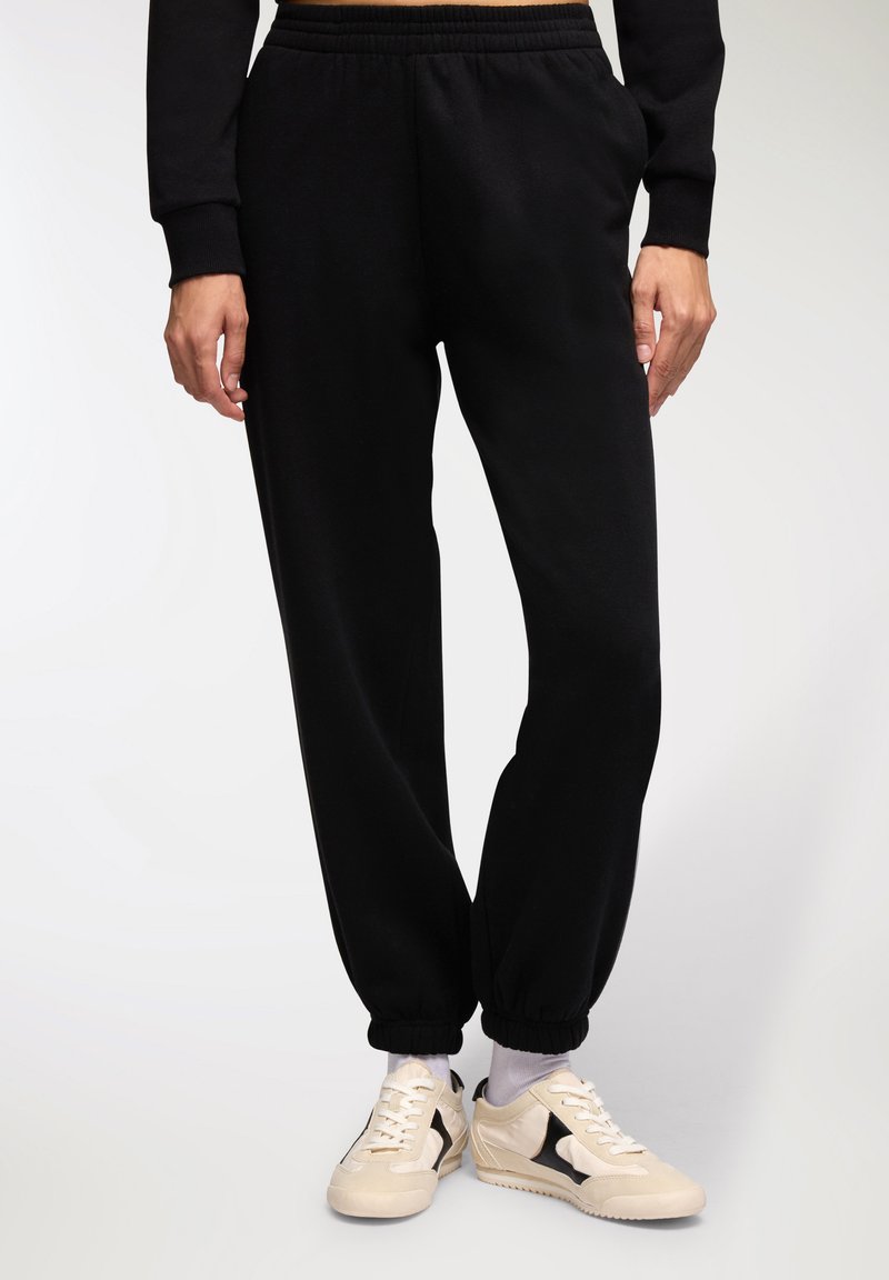 Pantaloni della tuta neri con cintura elastica e polsini a chiusura stretta, realizzati in materiale morbido. Abbinati a sneakers crema con dettagli neri.